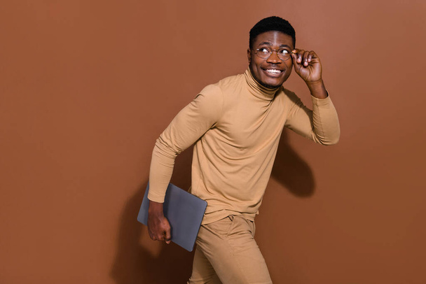 Portrait of attractive cheerful trendy guy holding laptop walking touching specs deciding isolated over brown color background. - Foto, Imagem