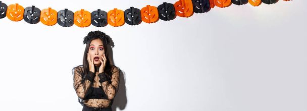 Bild einer Frau im Hexenkostüm, die erschrocken schaut, Schrecken oder Angst ausdrückt, während sie über weißem Hintergrund mit Kürbisfahnen-Dekoration steht und Halloween feiert. - Foto, Bild