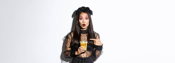 Erstauntes asiatisches Mädchen blickt in die Kamera, während es mit dem Finger auf Süßigkeiten zeigt, sammelt Leckereien zu Halloween, trägt Hexenkleid, steht über weißem Hintergrund. - Foto, Bild