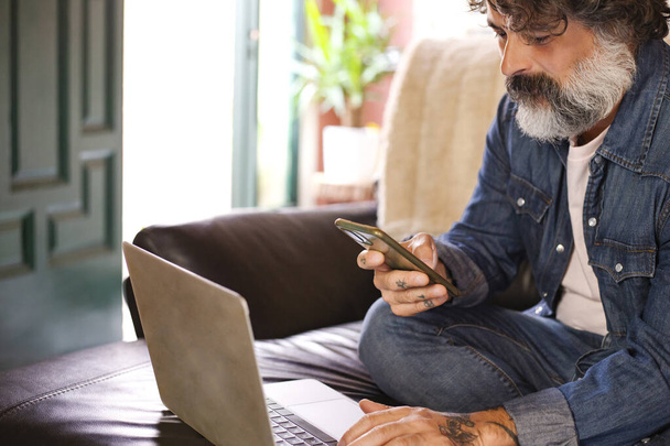 Valkoihoinen keski-ikäinen parrakas mies käyttää kannettavaa tietokonetta ja älypuhelinta, etätyötä, online-sosiaalista mediaa kotona - Valokuva, kuva