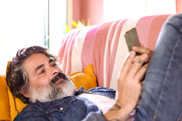 Szczęśliwy uśmiechnięty mężczyzna korzystający ze smartfona siedząc na kanapie w domu. Dojrzały mężczyzna leżący na kanapie czytając wiadomości w telefonie komórkowym. - Zdjęcie, obraz