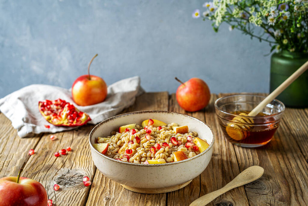 Sweet homemade barley porridge in bowl with pomegranate seeds, apples and honey on wooden table with ingredients and wild flowers. Rosh Hashanah celebration concept. - Photo, image