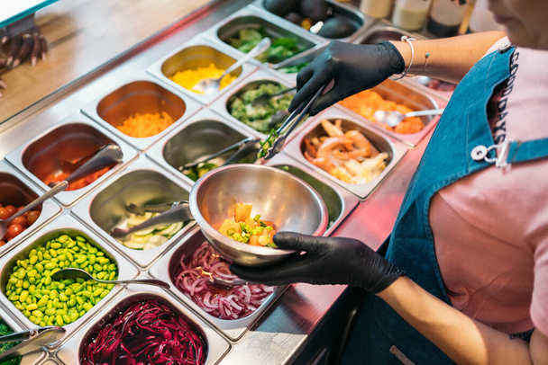 Unrecognized restaurant worker in protective gloves adding ingredients into poke bowl. - Foto, Bild