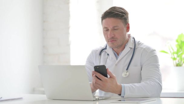 Середній літній доктор користується смартфоном під час роботи з ноутбуком в офісі. - Фото, зображення