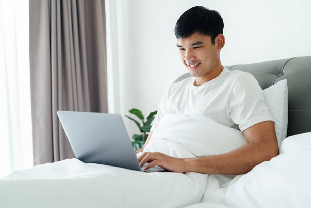 Asiatique homme en blanc t-shirt pose sur le lit en utilisant ordinateur portable dans la chambre. - Photo, image
