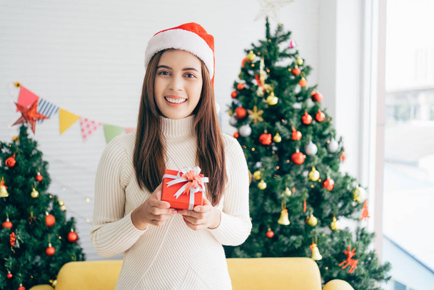 Молода азіатка, одягнена в светр і капелюх Санта-Клауса, посміхається і тримає різдвяний подарунок у червоній коробці з стрічкою у вітальні з прикрашеною ялинкою на задньому плані.. - Фото, зображення