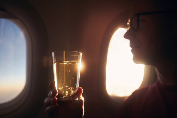 Man enjoying drink during flight. Passenger holding glass of sparkling wine against airplane window at sunset - Photo, image