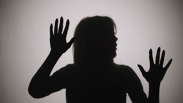 Black silhouette of a woman touching the glass with her palms and looking at the camera. The outline of a woman against the background of a window with white matte light. Model poses in studio with - Foto, Imagem