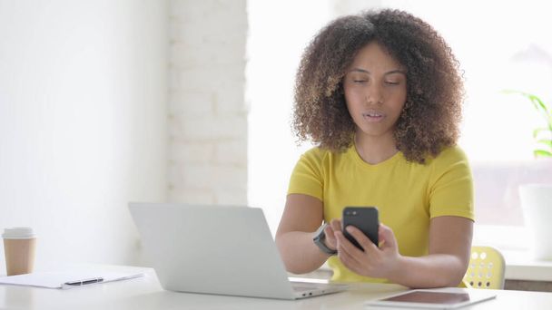 Jovem mulher africana usando Smartphone ao usar Laptop no escritório - Foto, Imagem