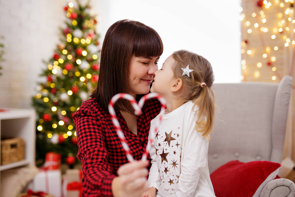 Cute mother and daughter touching noses in christmas decorated room. Heart from christmas candy in hand - Foto, Imagem