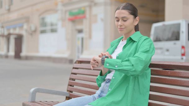 Молодая женщина использует умные часы, сидя на скамейке под открытым небом - Фото, изображение
