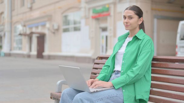 Відкидаючи молоду жінку у запереченні, використовуючи лаптоп сидячи надворі на лаві - Фото, зображення