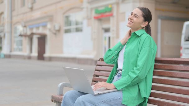 Young Woman with Neck Pain Using Laptop while Sitting Outdoor on Bench - 写真・画像