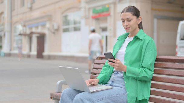 Young Woman Using Smartphone and Laptop while Sitting Outdoor on Bench - Fotografie, Obrázek