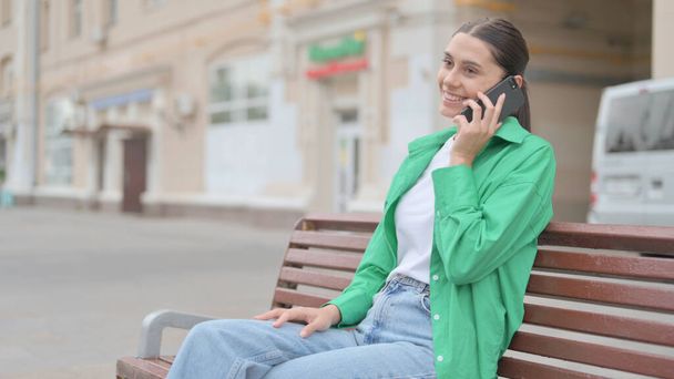 Молодая женщина разговаривает по телефону, сидя на скамейке - Фото, изображение