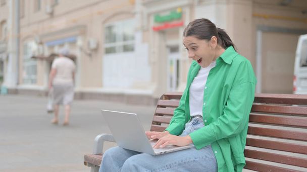 Молодая женщина празднует успех на ноутбуке, сидя на скамейке - Фото, изображение