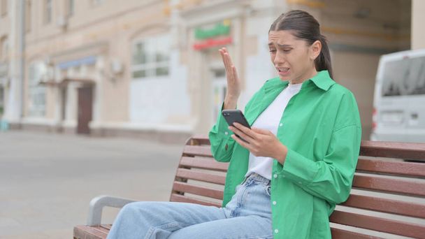 Молодая женщина реагирует на потерю на смартфоне, сидя на скамейке запасных - Фото, изображение