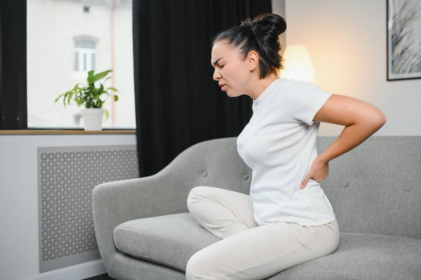 Upset mature woman suffering from backache, unhappy senior blonde sitting on a sofa at living room, feeling discomfort because of pain in back. - Фото, изображение