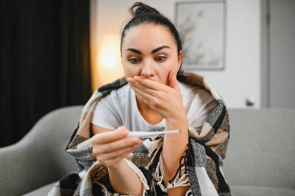 Cold and flu. Sick woman caught cold, feeling illness. Closeup of beautiful unhealthy girl covered in blanket wiping nose and looking at thermometer. Healthcare concept - 写真・画像