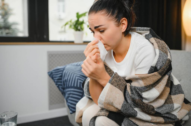 Sick woman with a headache sitting on a sofa at home wrapped in grey blanket. - Zdjęcie, obraz