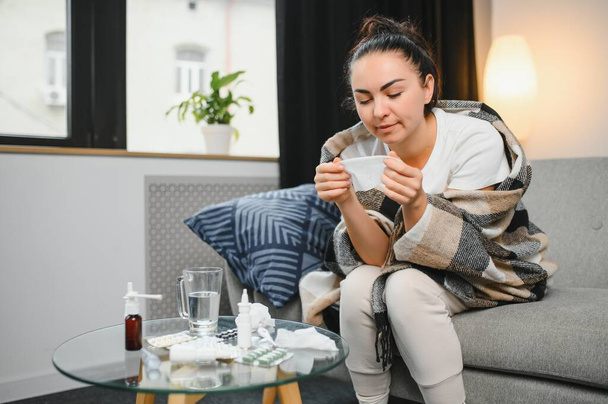Ill young woman sit on sofa covered with blanket, freezing, blowing her runny nose, sneeze in tissue. Female got fever, flu and influenza symptoms cough at home. Sick allergic with respiratory disease. - Foto, imagen