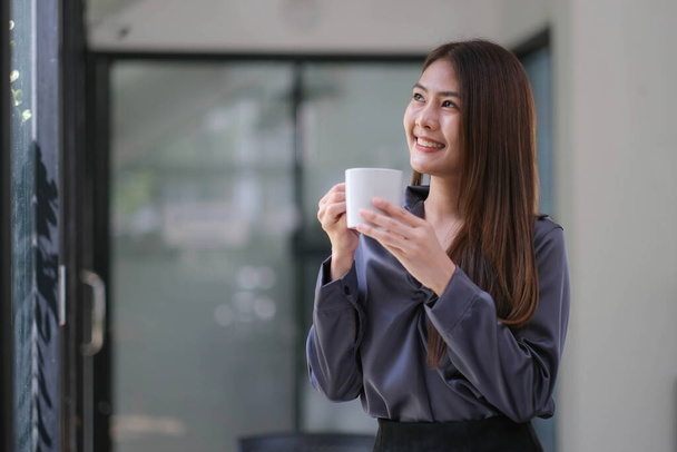 Magabiztos ázsiai üzletasszony, keresztbe tett karokkal áll egy kávéscsészével a kezében, mosolyog és a kamerába néz. - Fotó, kép