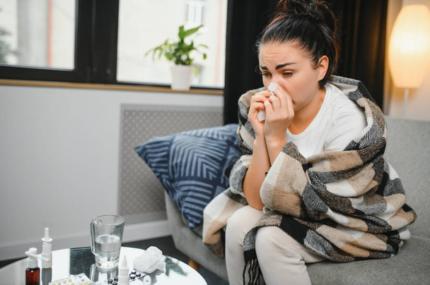 Ill young woman sit on sofa covered with blanket, freezing, blowing her runny nose, sneeze in tissue. Female got fever, flu and influenza symptoms cough at home. Sick allergic with respiratory disease. - Valokuva, kuva