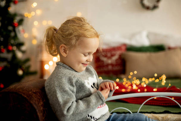 Linda niña rizada en suéter de Navidad de invierno, sentada en una habitación acogedora, decorada para Navidad - Foto, Imagen