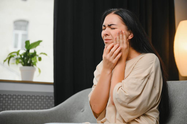 Woman with severe toothache touching her swollen cheek. Young girl feeling discomfort after tooth extraction. Upset lady with inflamed tooth nerve or terrible dental pain sitting on sofa at home - Photo, Image