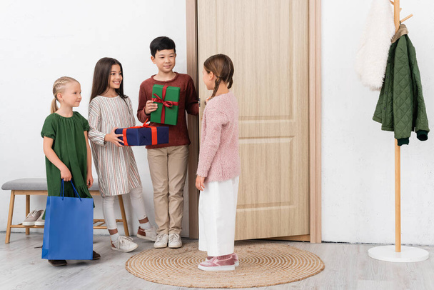 Smiling multiethnic kids with presents standing near friend and door in hallway at home  - Zdjęcie, obraz