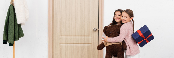 Chicas positivas con oso de peluche y presente abrazos en casa, pancarta  - Foto, Imagen