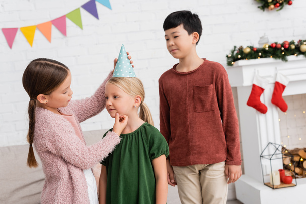 Girl wearing party cap on friend near asian boy during birthday party at home  - Foto, Imagen