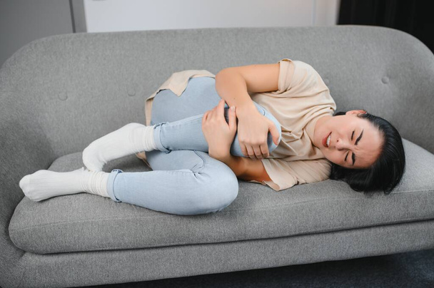 Beautiful hispanic woman in painful expression holding her belly suffering menstrual period pain lying sad on home bed having tummy cramp in female health concept. - Foto, imagen