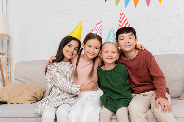 Smiling interracial kids in party caps looking at camera and hugging during birthday party at home - Фото, зображення
