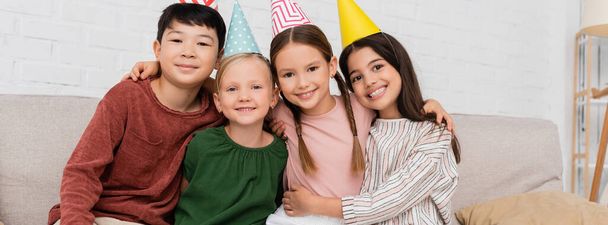 Feliz interracial niños en gorras de fiesta mirando a la cámara durante la fiesta de cumpleaños en casa, pancarta  - Foto, Imagen