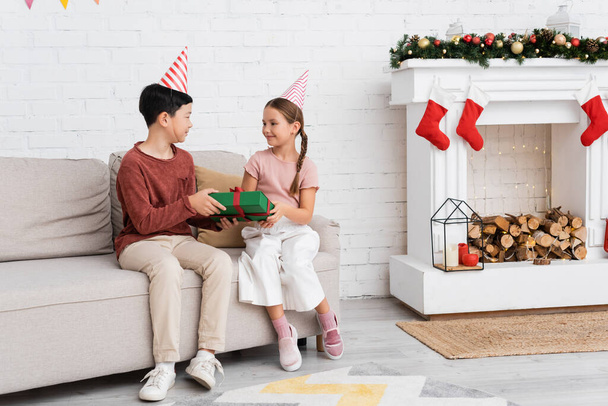 Asian boy in party cap holding present near friend on couch and christmas decor at home  - Foto, afbeelding