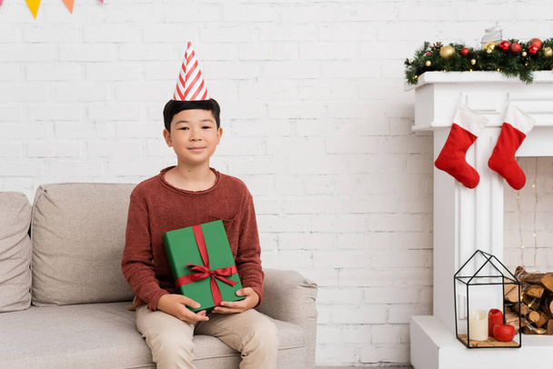 Preteen asian boy in party cap holding present on couch near christmas decor at home  - 写真・画像