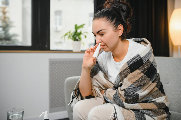 Nasal spray in hand of sick young woman sitting on couch. Allergic rhinitis symptoms and treatment. - Fotoğraf, Görsel