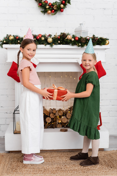 Lächelnde Kinder in Partymützen mit Geschenk und Blick in die Kamera am Kamin mit Weihnachtsstrümpfen zu Hause  - Foto, Bild