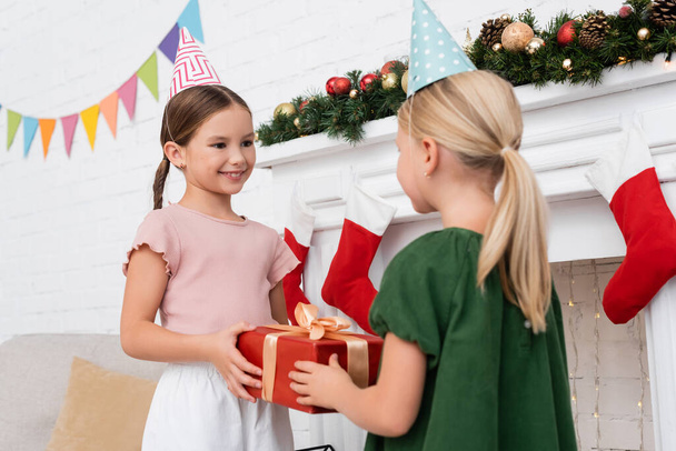 Smiling girl in party cap taking present from blurred friend near christmas decor at home  - Фото, изображение