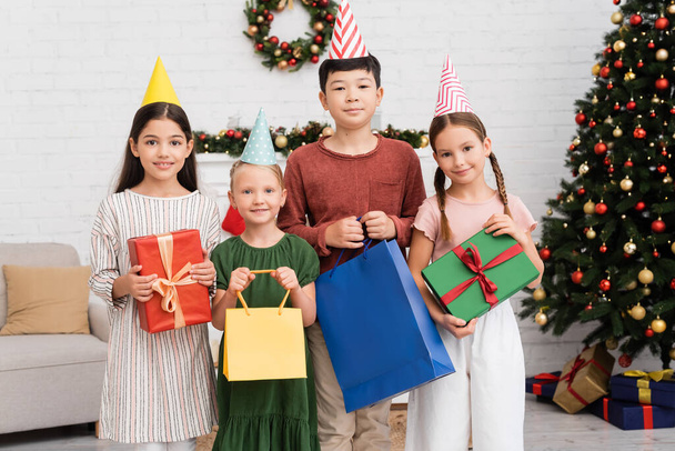 Crianças multiétnicas em bonés de festa segurando sacos de compras e presentes perto de decoração de Natal borrada em casa  - Foto, Imagem