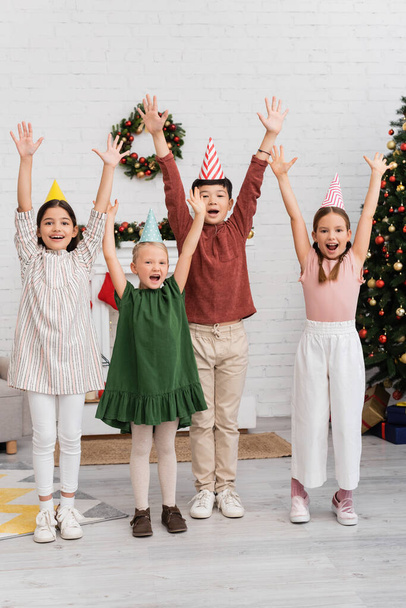 Cheerful interracial kids in party caps celebrating birthday party near blurred christmas decor at home  - Photo, Image
