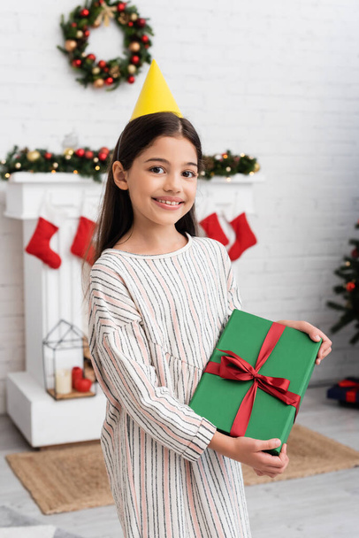 Portrait of cheerful girl in party cap holding gift box during birthday party in winter  - Foto, Bild