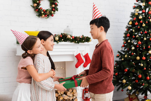 Asian boy in party cap giving present to cheerful friend near christmas decor on fireplace at home  - Photo, Image