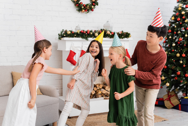 Alegre interracial niños en partido caps jugando etiqueta durante cumpleaños fiesta cerca de Navidad decoración en casa  - Foto, Imagen