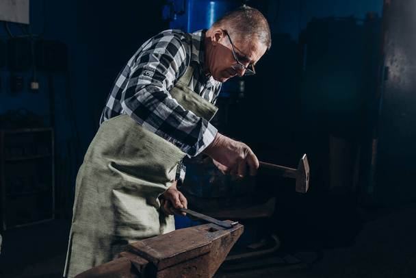 Senior an artisan blacksmith knocks with a hammer on iron to shape. - 写真・画像