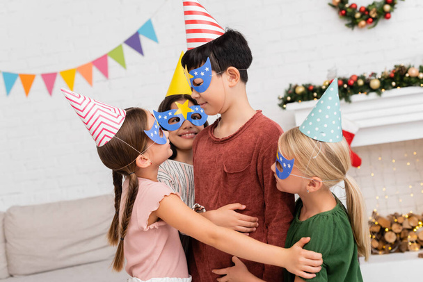 Smiling kids hugging asian friend in party cap and mask during birthday celebration at home  - Fotografie, Obrázek