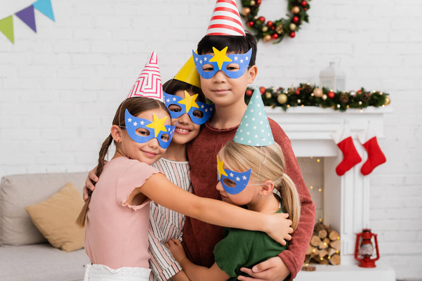 Allegri bambini interrazziali in berretti e maschere da festa che si abbracciano durante la festa di compleanno a casa  - Foto, immagini