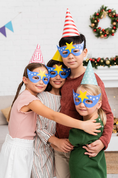 Positive interrassische Kinder in Partymützen und Masken, die sich zu Hause umarmen und in die Kamera schauen  - Foto, Bild