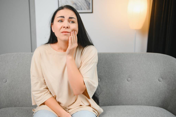 Woman with severe toothache touching her swollen cheek. Young girl feeling discomfort after tooth extraction. Upset lady with inflamed tooth nerve or terrible dental pain sitting on sofa at home - Photo, Image